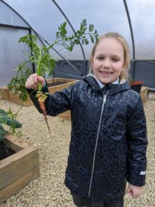 A young girl is seen wearing her winter coat and holding up a freshly picked Carrot for the camera in the academy garden area.