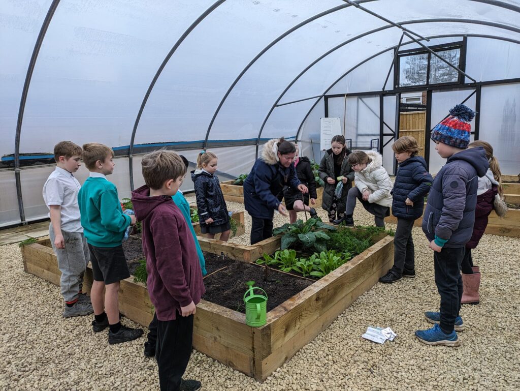 A group of pupils are pictured gathered round a member of staff, instructing them on how to garden inside a large greenhouse.