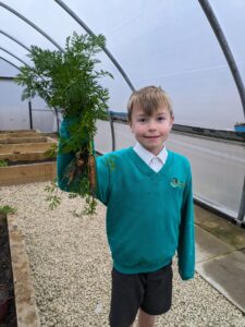 A young boy is shown wearing his academy uniform and smiling for the camera, whilst holding up a Carrot he has just picked in the academy greenhouse.