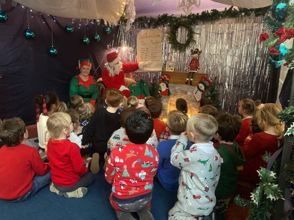 Photo showing Horsmonden pupils sat on the floor in Santa's Grotto, listening to him speak to them. Santa is seen pointing to an ornament on his mantelpiece.