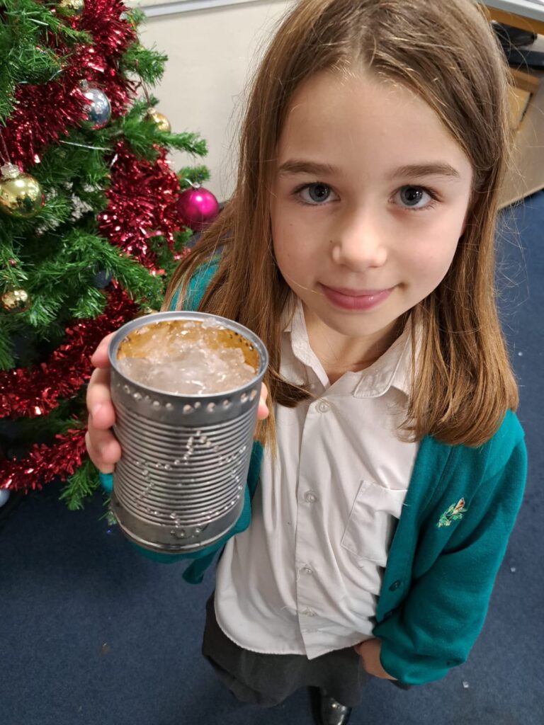A young girl in academy uniform is seen smiling for the camera, holding up a tin can she has made into a Christmas lantern.