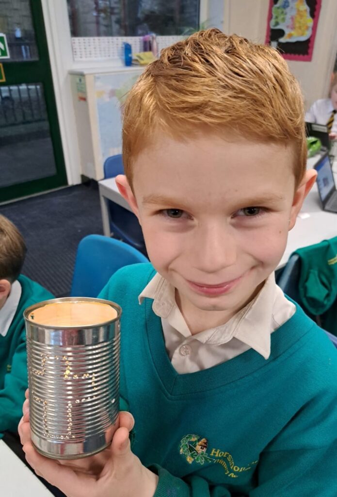 A young boy in academy uniform is seen smiling for the camera, holding up a tin can he has made into a Christmas lantern.