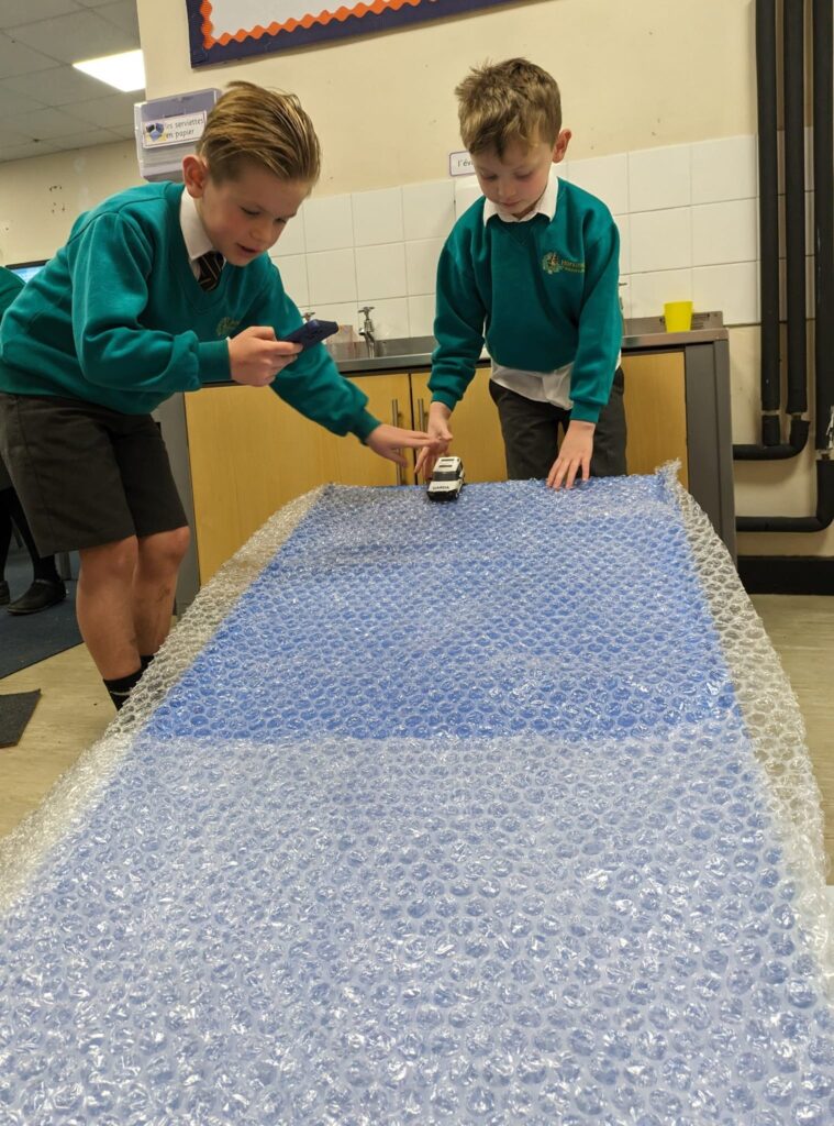 Two Year 6 pupils are pictured experimenting with friction by pulling a toy car across a sheet of bubble wrap.