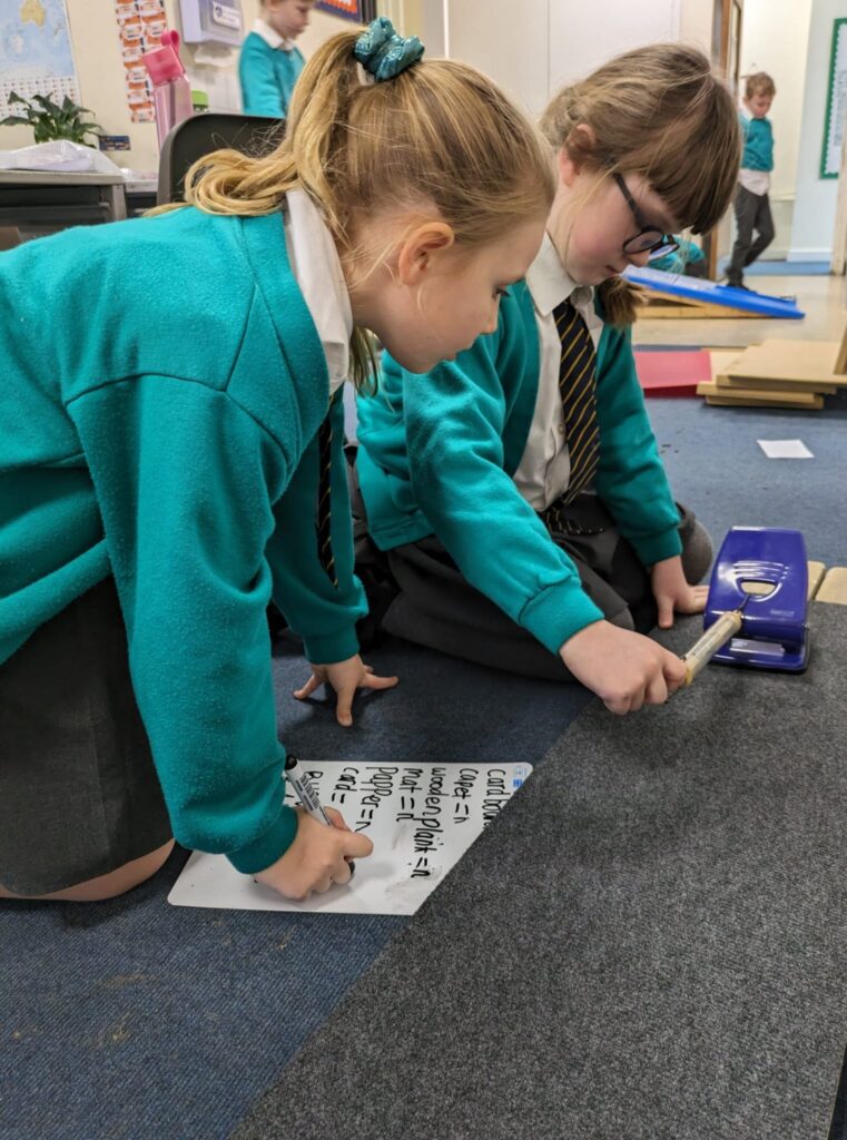 Two girls in Year 6 are seen experimenting with friction by pulling a holepunch along the carpet.