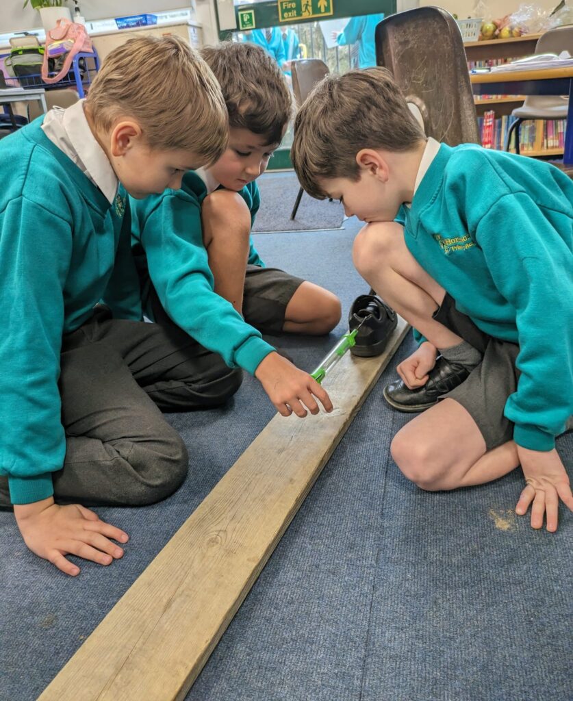 Three boys in Year 6 can be seen experimenting with friction by dragging an object along a long plank of wood.