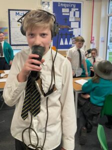 A young boy is seen standing in the middle of his classroom, trying on different items from WWII, such as goggles and headphones used by a pilot.
