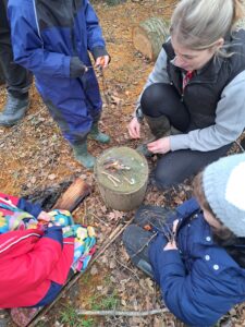 A member of staff is seen kneeling down to start a campfire in the Forest School. Three Year 1 pupils are pictured sat beside her.