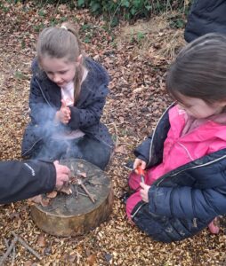 Two young girls from Year 1 are pictured sat around a small campfire in the Forest School, under the supervision of an adult member of staff.