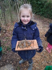A young girl in Year 1 is pictured smiling for the camera in the Forest School, whilst wearing her winter coat and holding a wooden tray with pine cones and twigs on it.