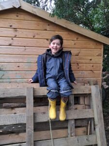 A young boy in Year 1 is pictured smiling for the camera, whilst sat on a wooden fence and wearing his winter coat in the Forest School.