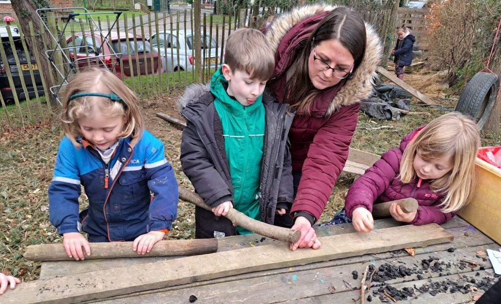 Three young pupils from Year 1 are seen interacting with some tree branches they have found in the Forest School, whilst under the supervision of an adult member of staff.