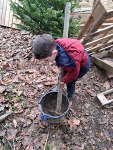 A young boy in Year 1 can be seen using a long tree branch to mix some mud in a plastic bucket in the Forest School.