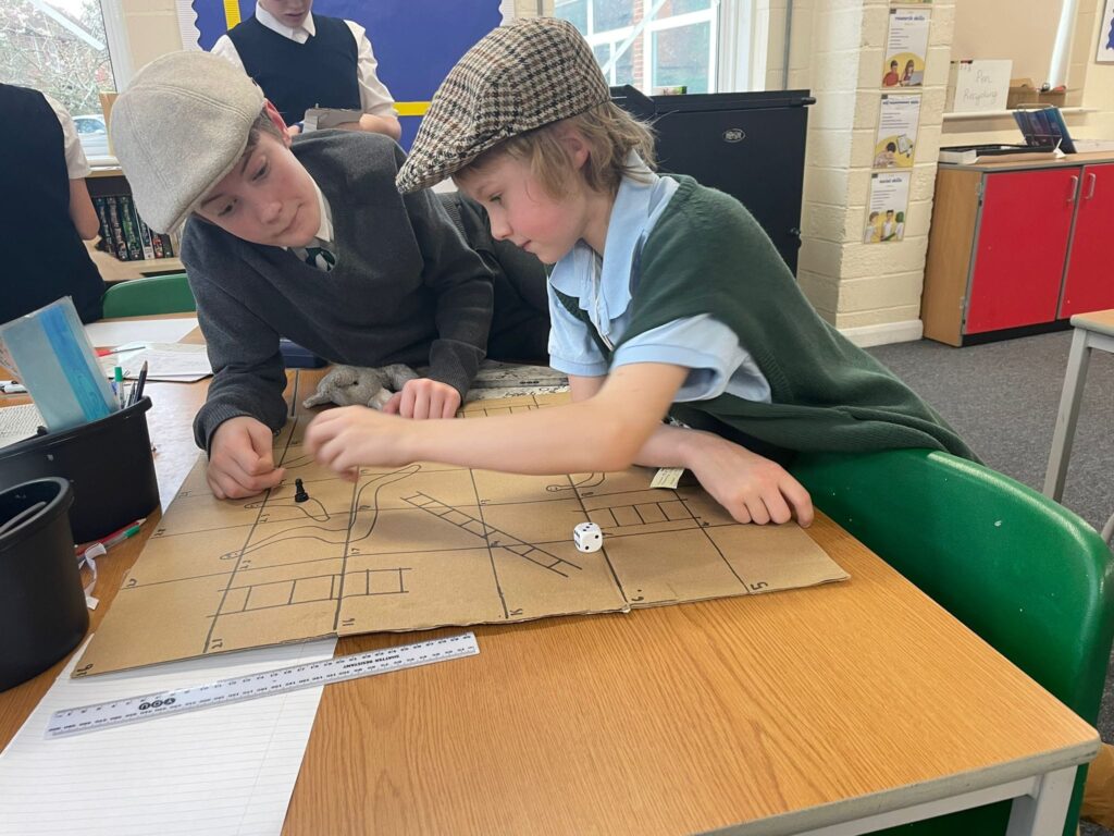 Students dressed as evacuees seen working together at a table to create a board for a game of 'Snakes & Ladders.'