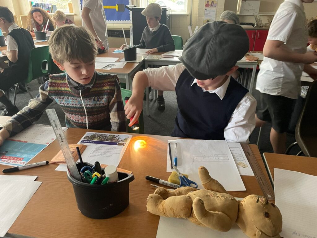Two male students are pictured sat together at a desk, wearing WWII evacuee clothing, looking at some items from that era.