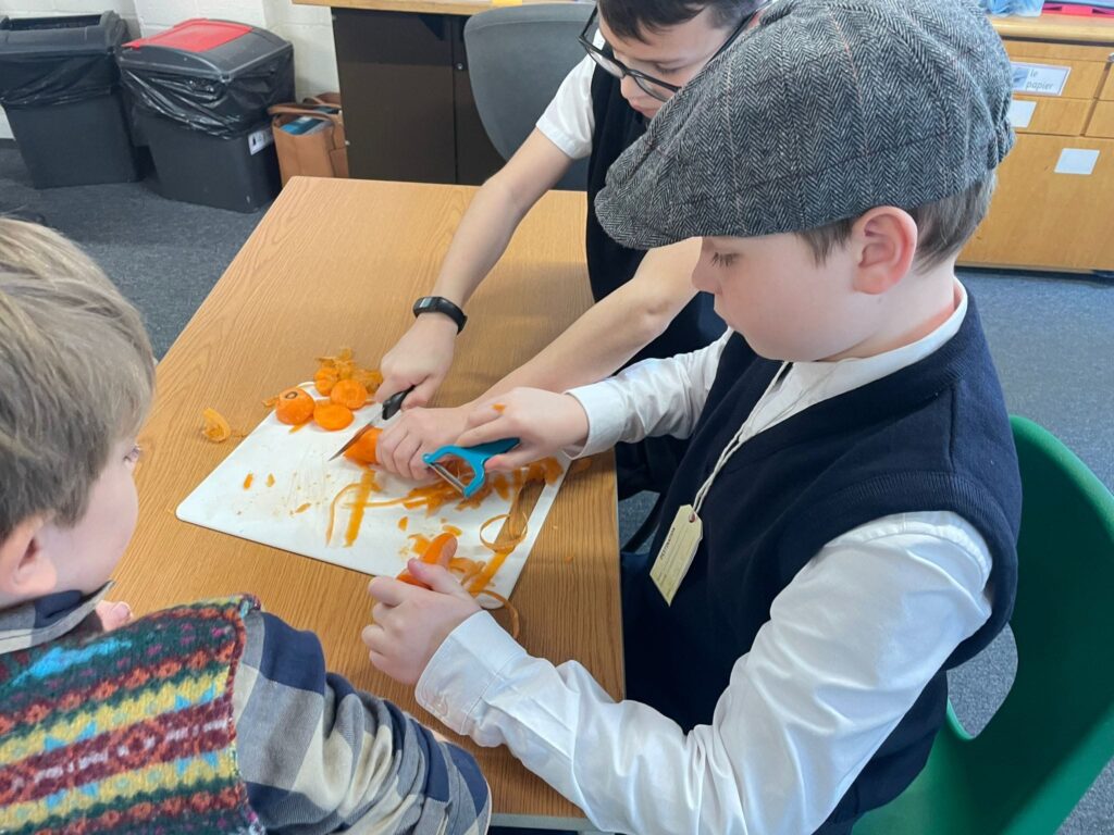 Three male students are pictured sat together at a desk, wearing WWII evacuee-style clothing, peeling carrots together.