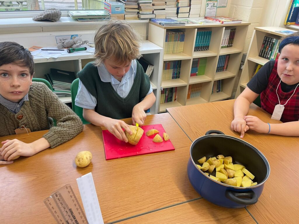 Three Year 6 students are pictured wearing WWII evacuee-style clothing, as they cut potatoes together at their desks.