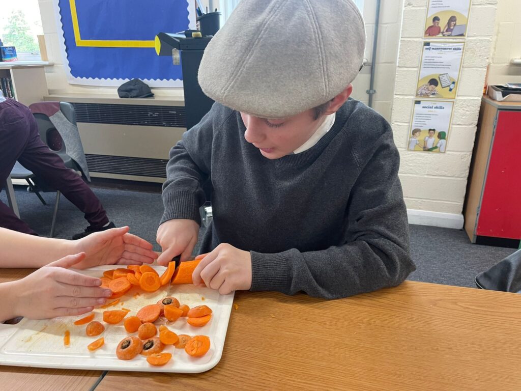 A Year 6 student can be seen sitting at his desk, cutting carrots, whilst wearing WWII evacuee-style clothing.