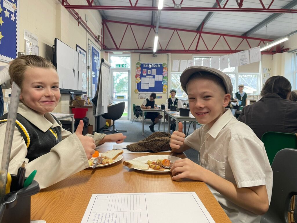 Two Year 6 students, a boy and a girl, can be seen sat at their desks, wearing WWII evacuee-style clothing and eating a meal.
