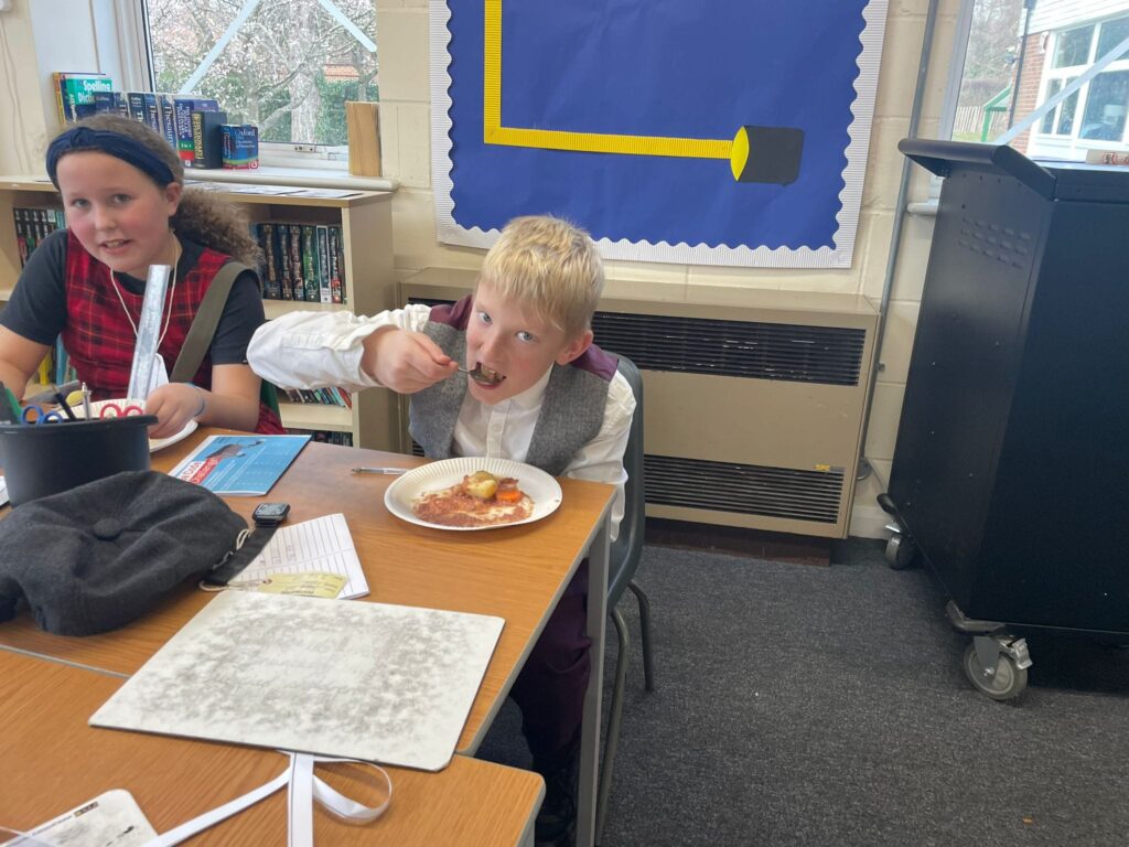 Two Year 6 students, a boy and a girl, are pictured sat next to one another, eating lunch. They are both dressed in WWII evacuee-style clothing.