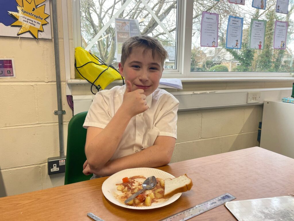 A male Year 6 student can be seen smiling for the camera with his thumb up, whilst eating his lunch.