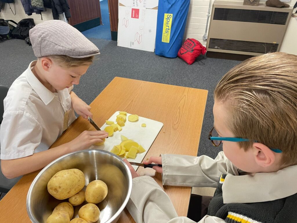 Two male Year 6 students are pictured sat together at a desk, chopping potatoes. They are both wearing WWII evacuee-style clothing.