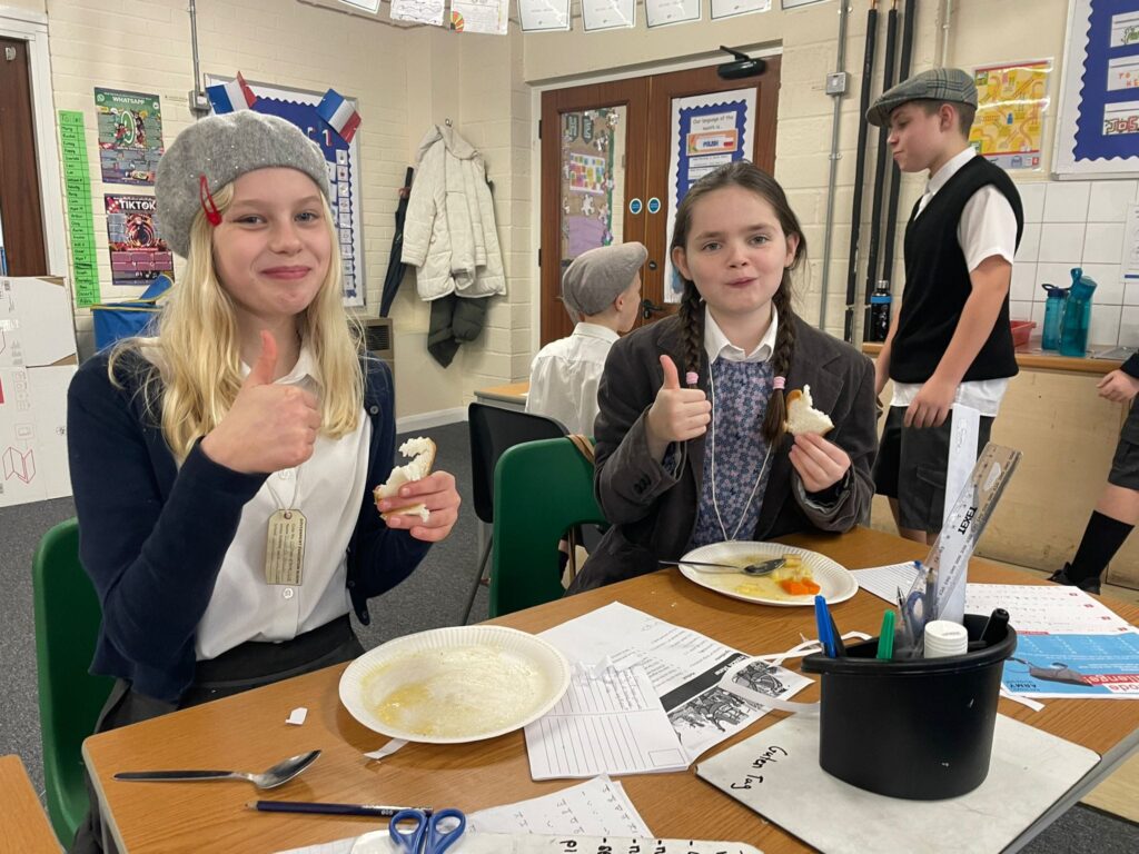 Two female Year 6 students are seen wearing WWII evacuee-style clothing and smiling for the camera, as they eat their lunch.